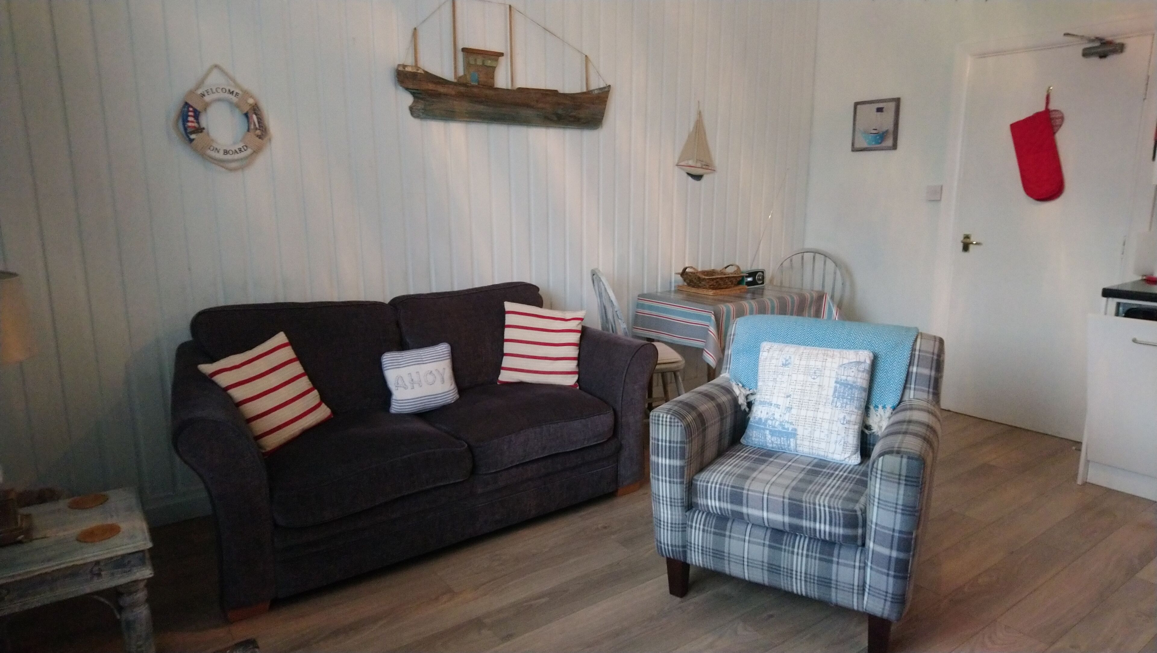 A living room with a dark sofa, a plaid armchair, and a small table with a radio. A ship model hangs on the wall, and a red stocking is hanging on the door.