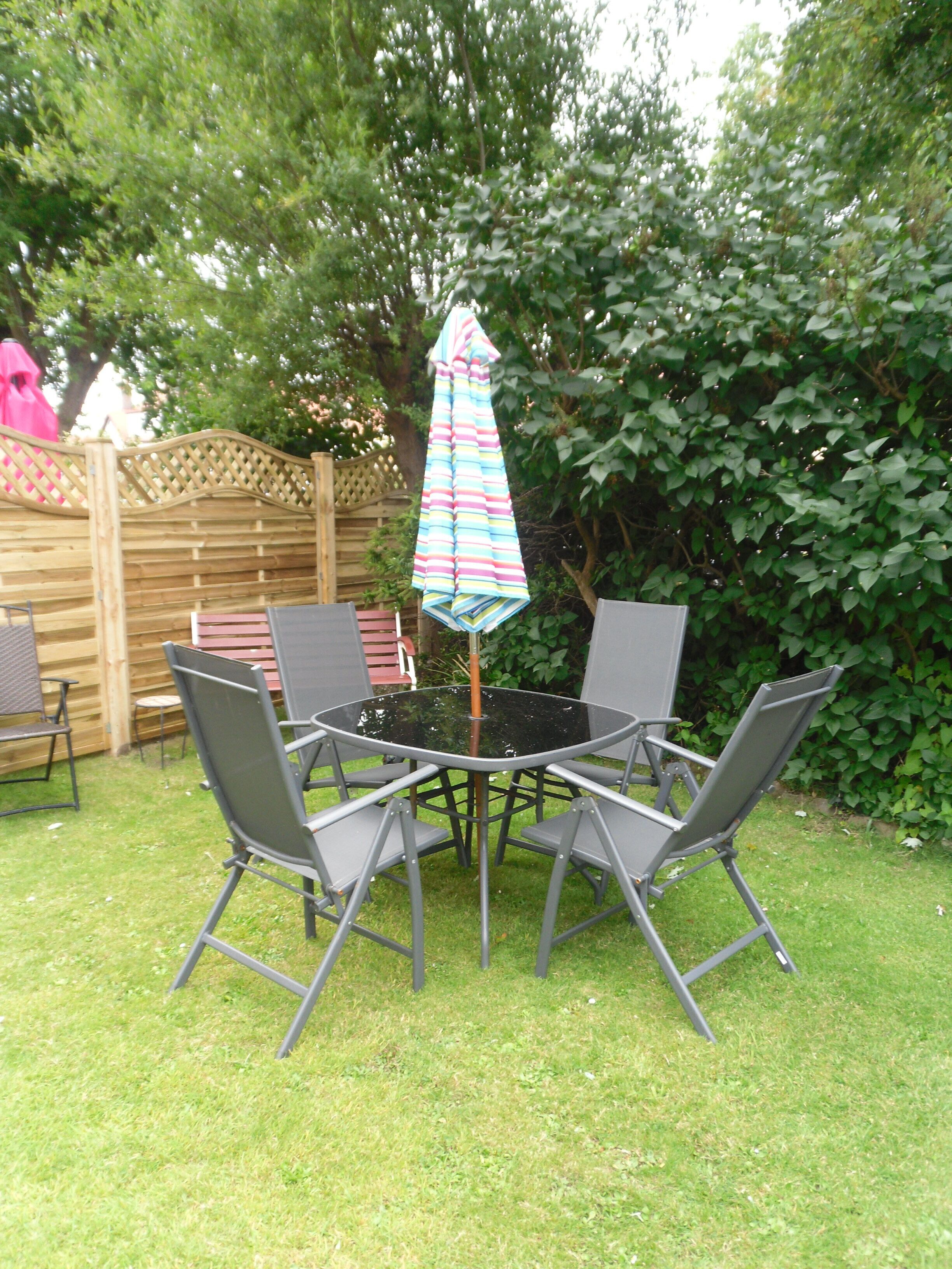 A round table with four grey chairs and a striped umbrella in a garden setting.