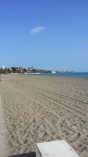 Une vaste plage de sable fin, parsemée d’empreintes de pas qui mènent à l’eau, un ciel bleu clair et des bâtiments visibles à l’arrière-plan.
