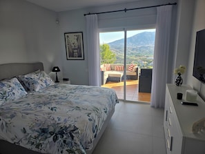 Une chambre à coucher comprenant un lit, une table de chevet, une lampe, une commode, un téléviseur et un balcon avec vue sur les montagnes.