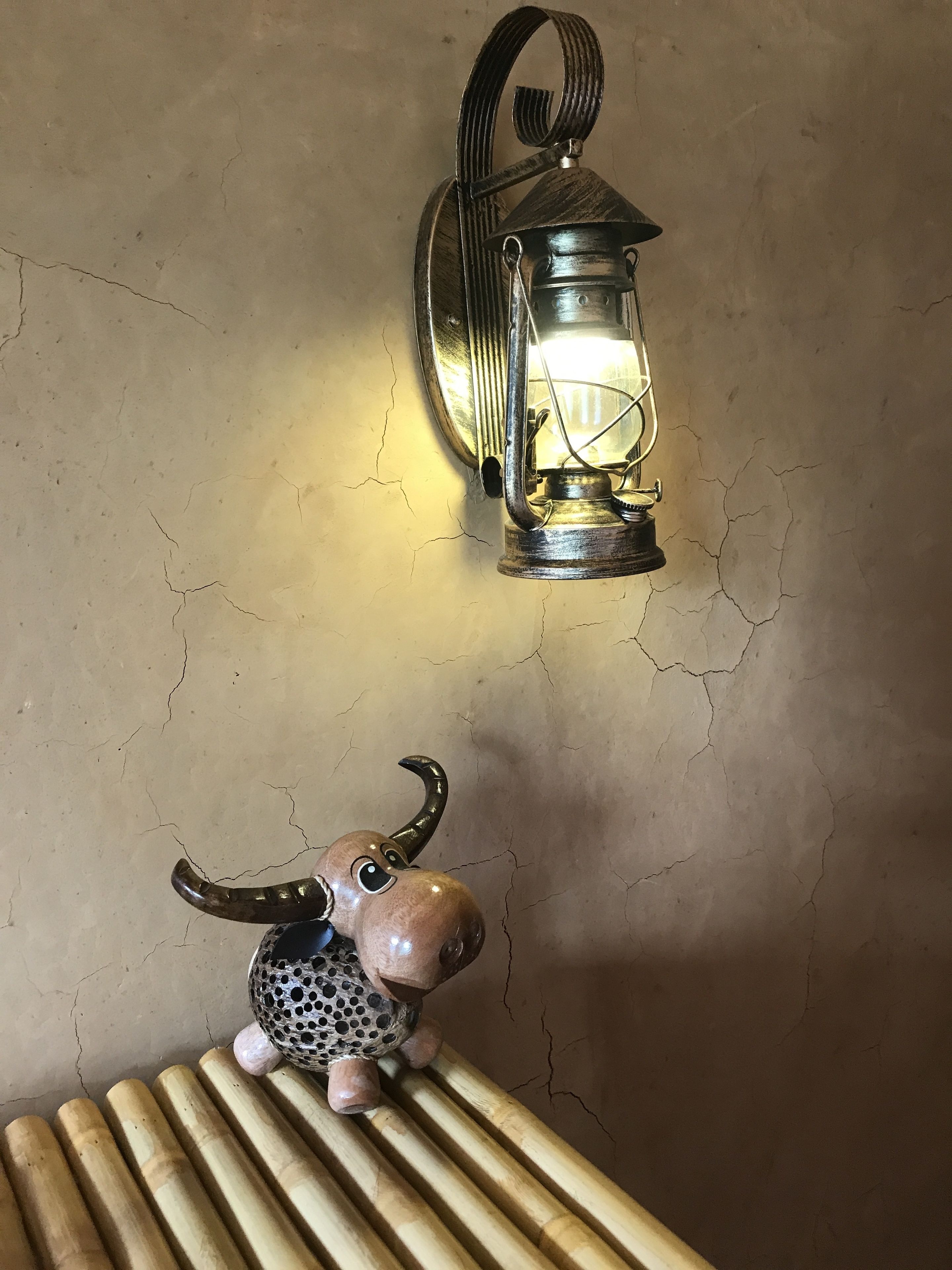 Una pared rústica con textura agrietada, un farol de metal con asa, y una pequeña figurina de burro sobre una tabla de madera.