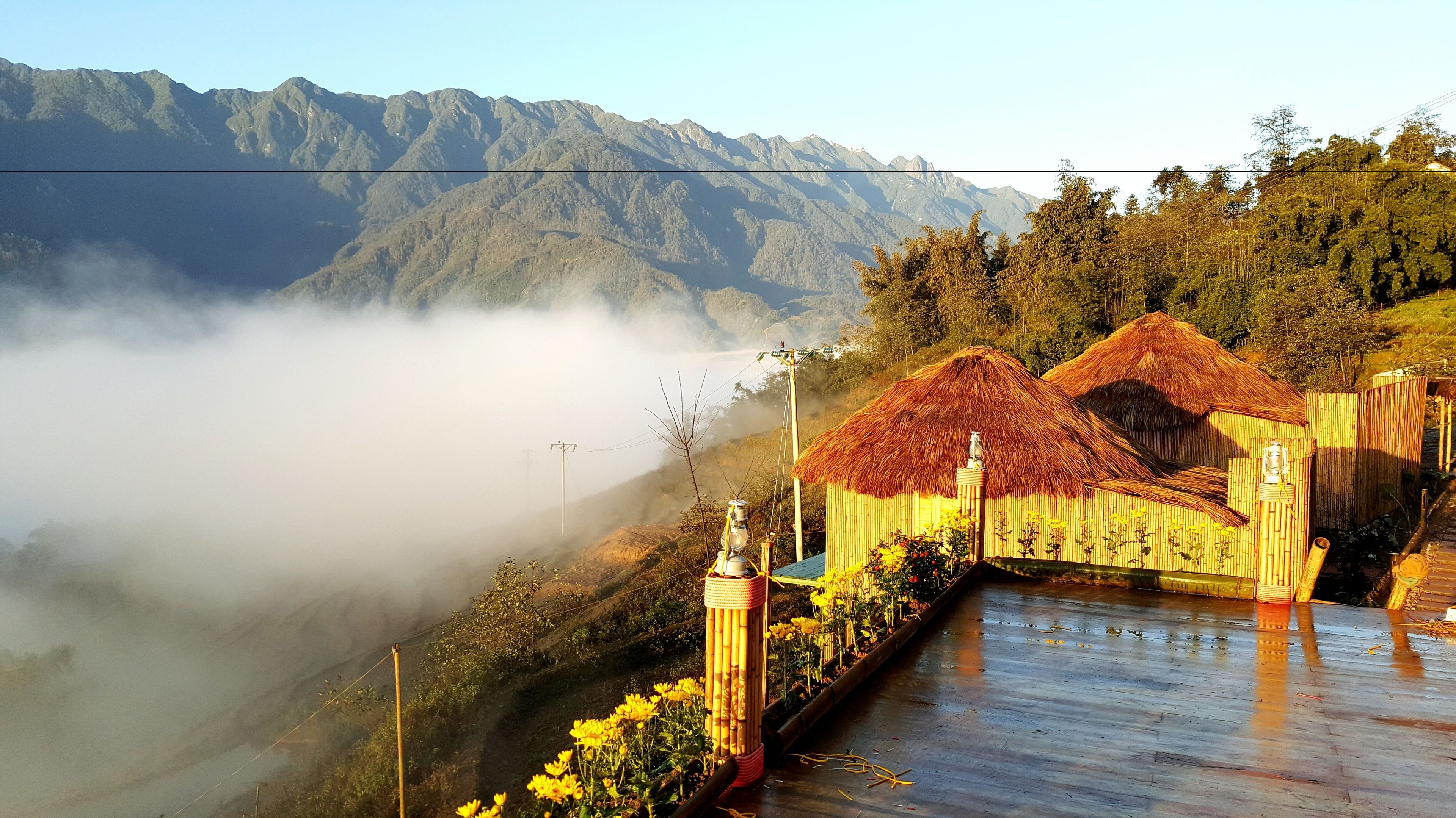 A traditional thatched-roof structure with a bamboo fence, set against a backdrop of mountains and mist.