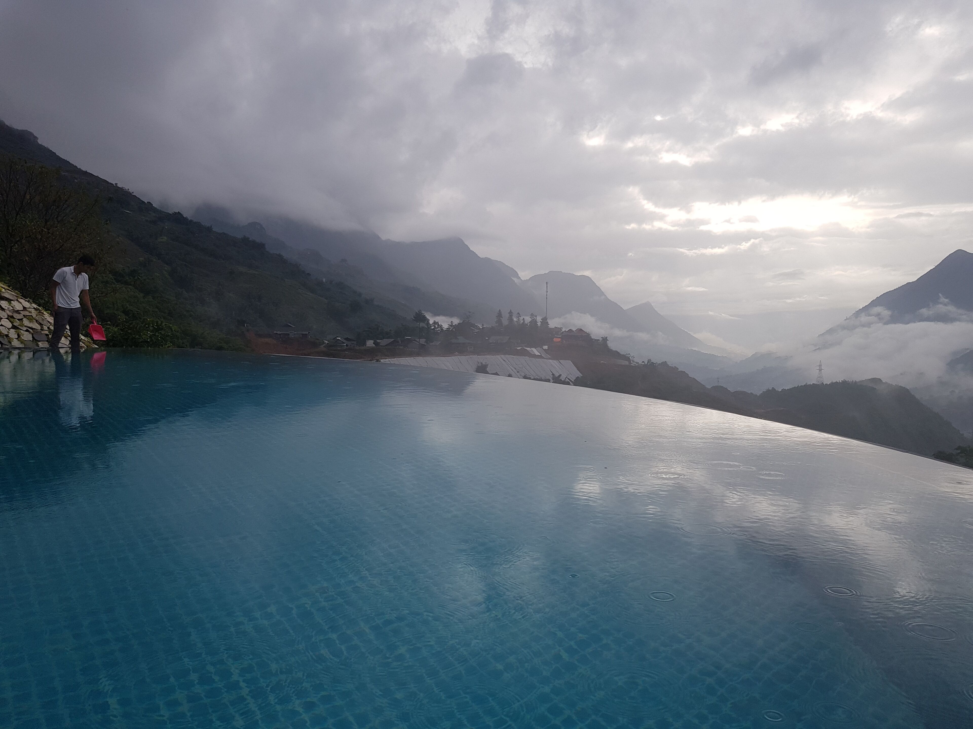 Una piscina azul cristalina con una persona de pie en el borde, rodeada de montañas y un cielo nublado.