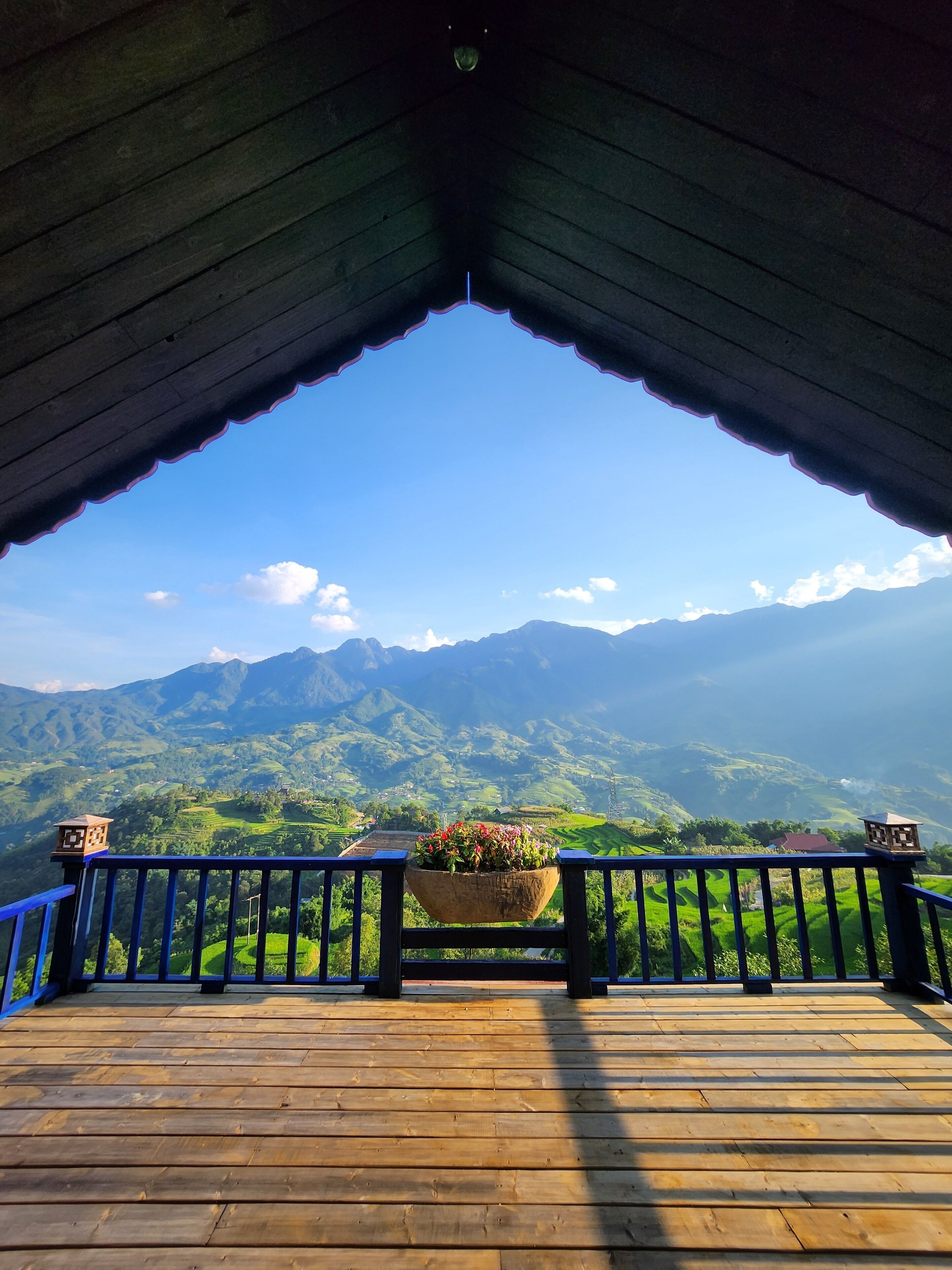 Une terrasse en bois avec une rambarde, un pot de fleurs et une vue sur des collines verdoyantes et des montagnes.
