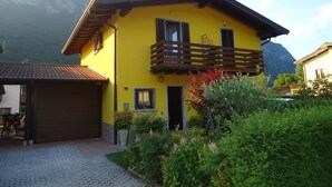 Ein gelbes, zweistöckiges Haus mit einem hölzernen Balkon, umgeben von viel Grün und Bergen im Hintergrund.