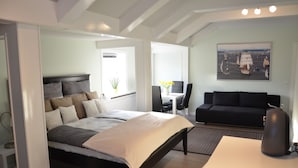 A modern bedroom with a large bed, a desk, a black sofa, and a painting of a sailing ship.
