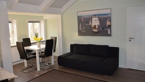 A modern living room with a black sofa, a dining table with chairs, and a large painting of sailboats on the wall.