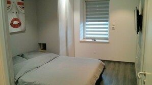 Un dormitorio con cama, ventana con persianas, puerta y una obra de arte colgada en la pared.