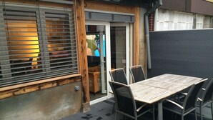 Un patio con mesa y sillas de madera, una puerta corrediza de vidrio y vista al jardín trasero.