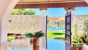 An outdoor dining area with a round table and wicker chairs, a pergola overhead, and a view of a pool and garden.