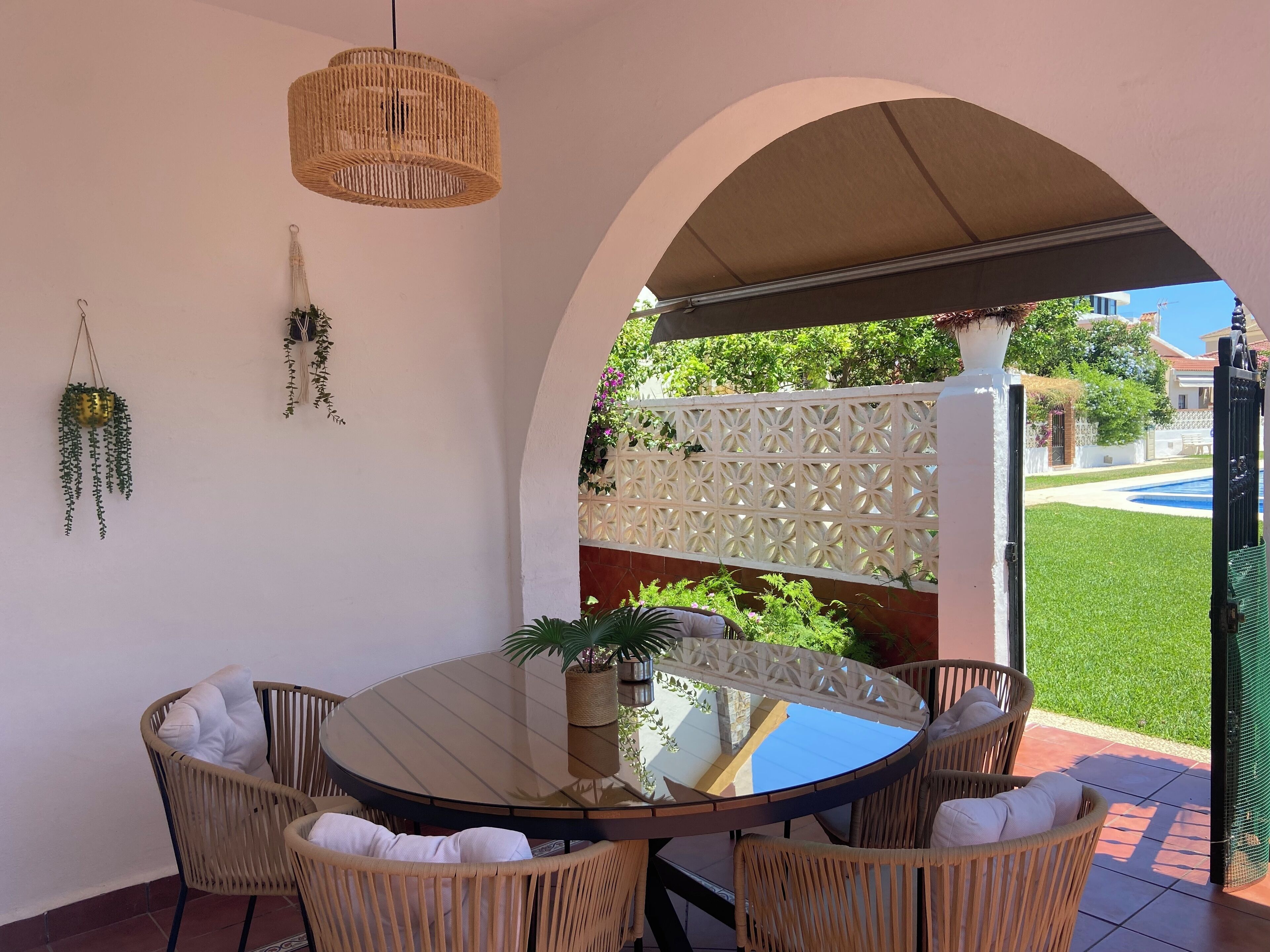 Un patio exterior cubierto con una mesa redonda y sillas de mimbre, un colgante de luz y vistas a un césped verde y edificios al fondo.