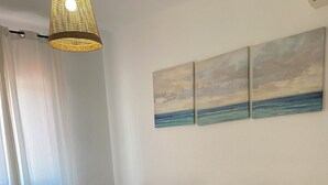 A neatly made bed with white linens and rolled towels, a bedside table with a lamp, and a wall with three paintings of a seascape.
