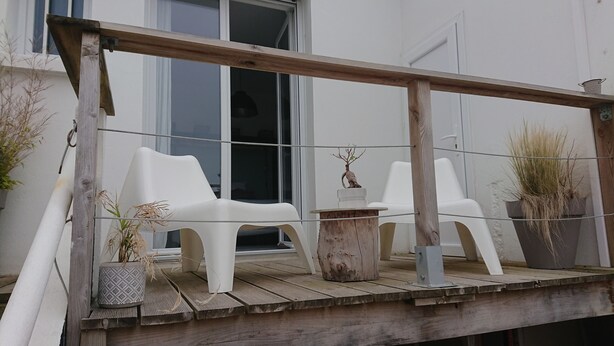 Una terraza de madera con dos sillas de plástico blancas y una pequeña mesa, rodeada de plantas en macetas.