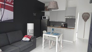 Una sala moderna con un sofá gris, una mesa blanca con un mantel individual azul y una zona de cocina con armarios blancos y un microondas.
