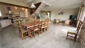 A spacious living area with a dining table set for six, surrounded by wooden chairs. A staircase with a stone accent wall leads to an upper level. - Cottage on the Crozon peninsula (Lanvéoc)