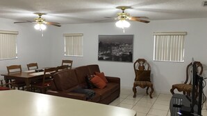 Un salon avec un canapé en cuir marron, une table à manger avec des chaises, deux ventilateurs de plafond et un grand tableau représentant un paysage urbain, encadré et accroché au mur.