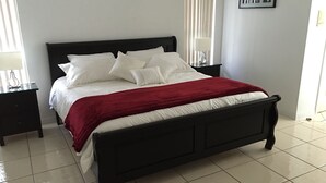 Un lit bien fait, avec du linge de lit blanc et une couverture rouge, flanqué de deux tables de chevet avec des lampes, dans une pièce aux murs blancs et au sol carrelé.