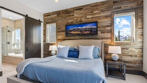 A modern bedroom with a large bed, wooden wall paneling, a mounted TV, and a bathroom with a glass shower.