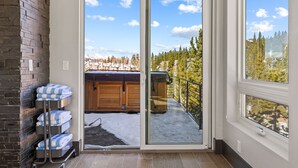 A room with a sliding glass door leading to a balcony with a hot tub and a view of a snowy mountain and trees.