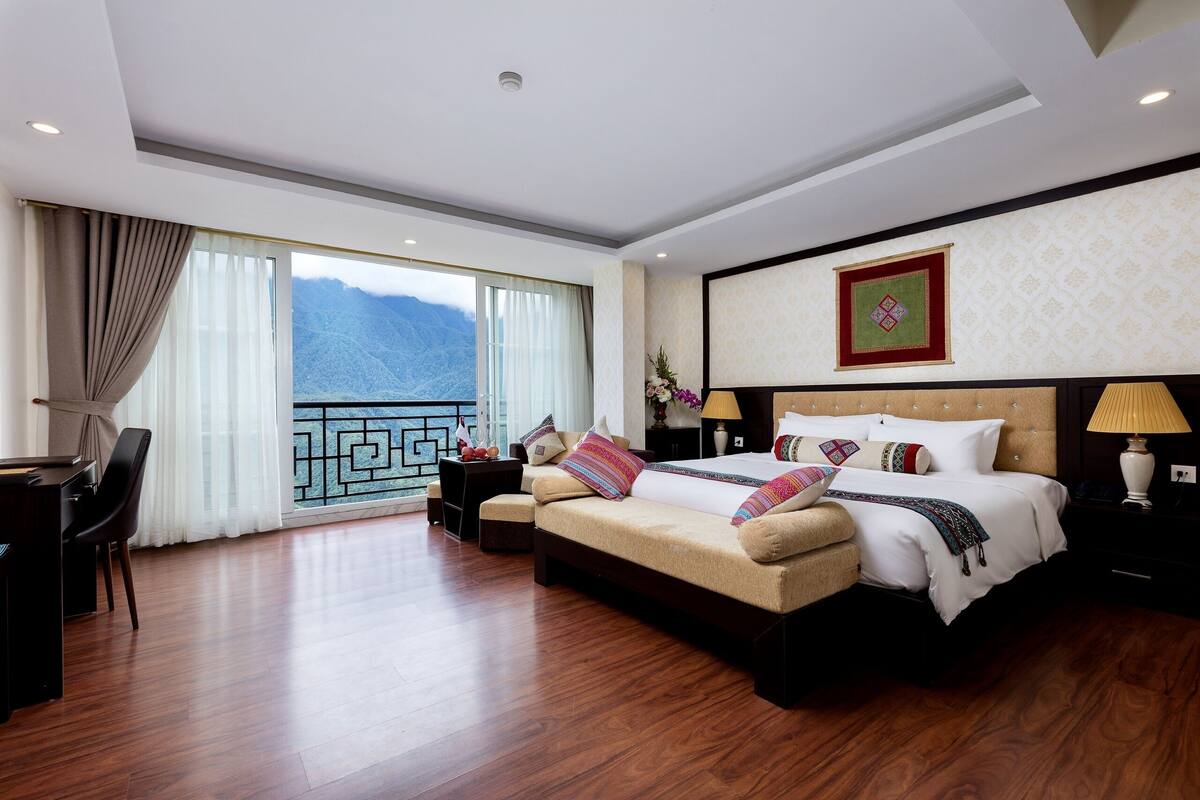 A spacious hotel room with a large bed, a desk, and a view of mountains through the window.