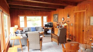 Interior acogedor de una cabaña de madera con sofá, sillones, televisor y comedor.