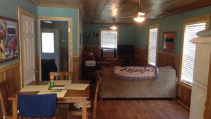A cozy room with a wooden ceiling, a dining table with chairs, a sofa with a 'Louisiana' blanket, a TV, and a window with blinds.