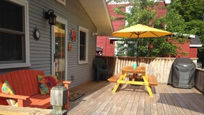Eine Holzterrasse mit Picknicktisch und Sonnenschirm, eine rote Couch mit bunten Kissen sowie ein Grill.