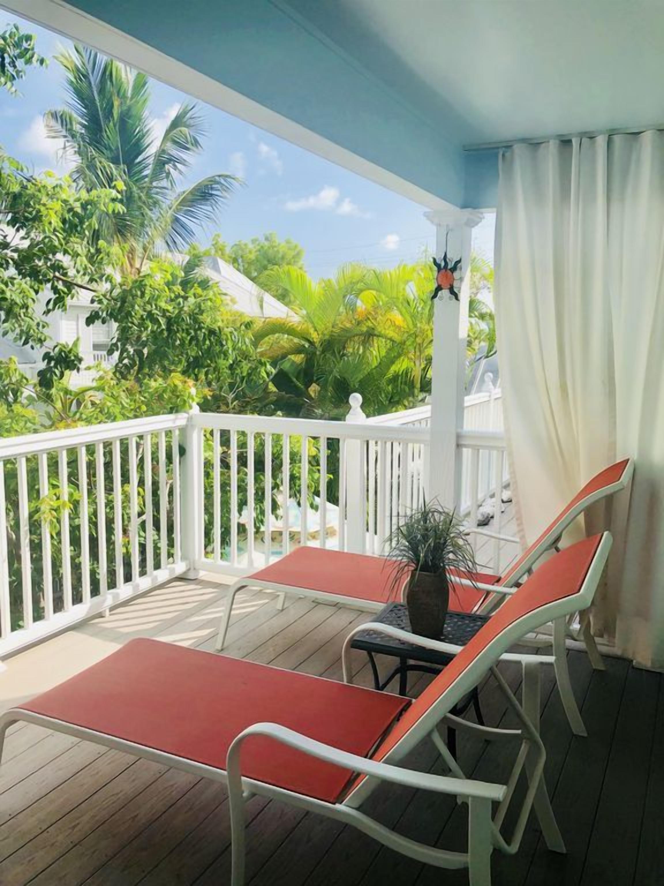 A covered outdoor seating area with a table and chairs, surrounded by a white railing, overlooking a lush garden with palm trees.