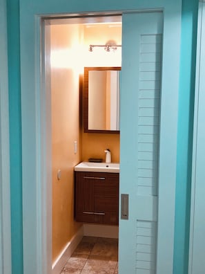 Een smalle gang die uitkomt bij een badkamer met een wastafel, spiegel en houten kasten.