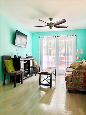 A living room with a wooden floor, a ceiling fan, a television mounted on the wall, a sofa, a chair, and a table.