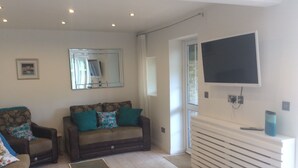 A living room with two sofas, a coffee table, a TV mounted on the wall, and a window with blinds. - Garden bungalow - dog friendly (Bushey)