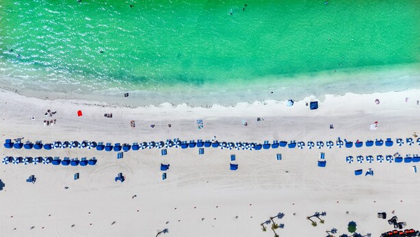 Una playa con hileras de sillas azules y sombrillas, una orilla arenosa y un mar turquesa transparente.