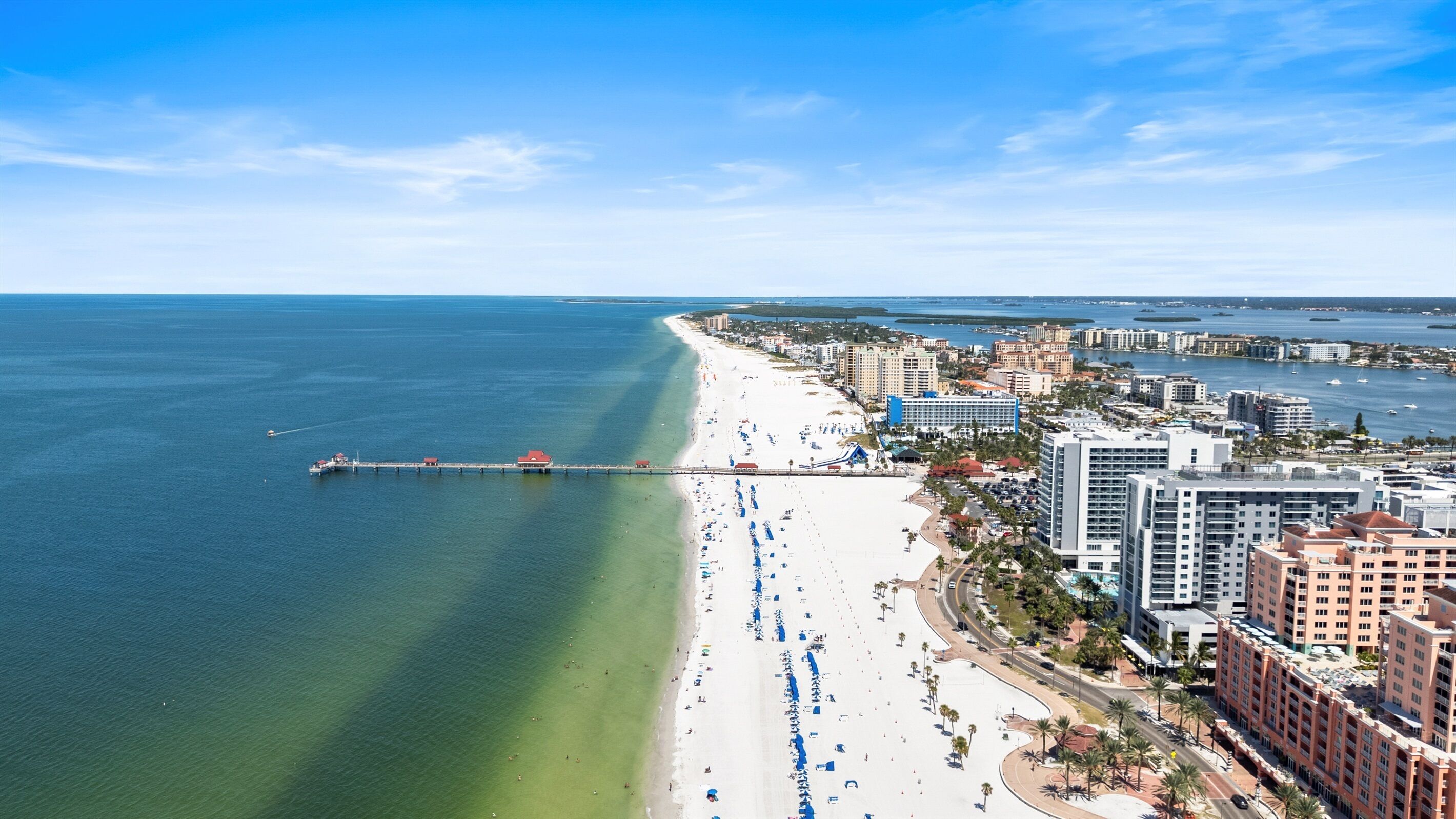A coastal cityscape with a long sandy beach, a pier extending into the sea, and high-rise buildings along the shore.