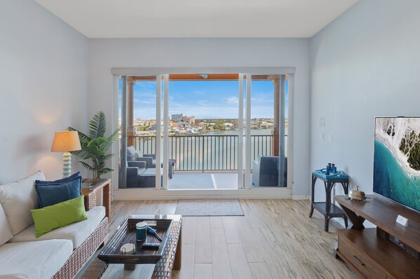 A modern living room with a balcony, a sofa with cushions, a coffee table, a TV, and a view of the city and water.