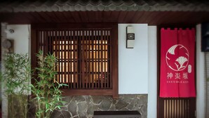 Uma entrada em estilo japonês tradicional, com janela de madeira entrelaçada, parede de pedra, planta de bambu em vaso, um lampião de pedra e uma faixa vermelha com caracteres japoneses.