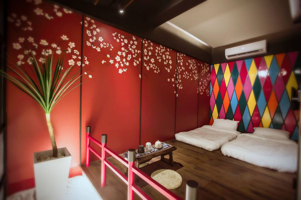 A room with a red wall, a colorful diamond-patterned wall, a white sofa, a small table with a plant, and a wooden floor.