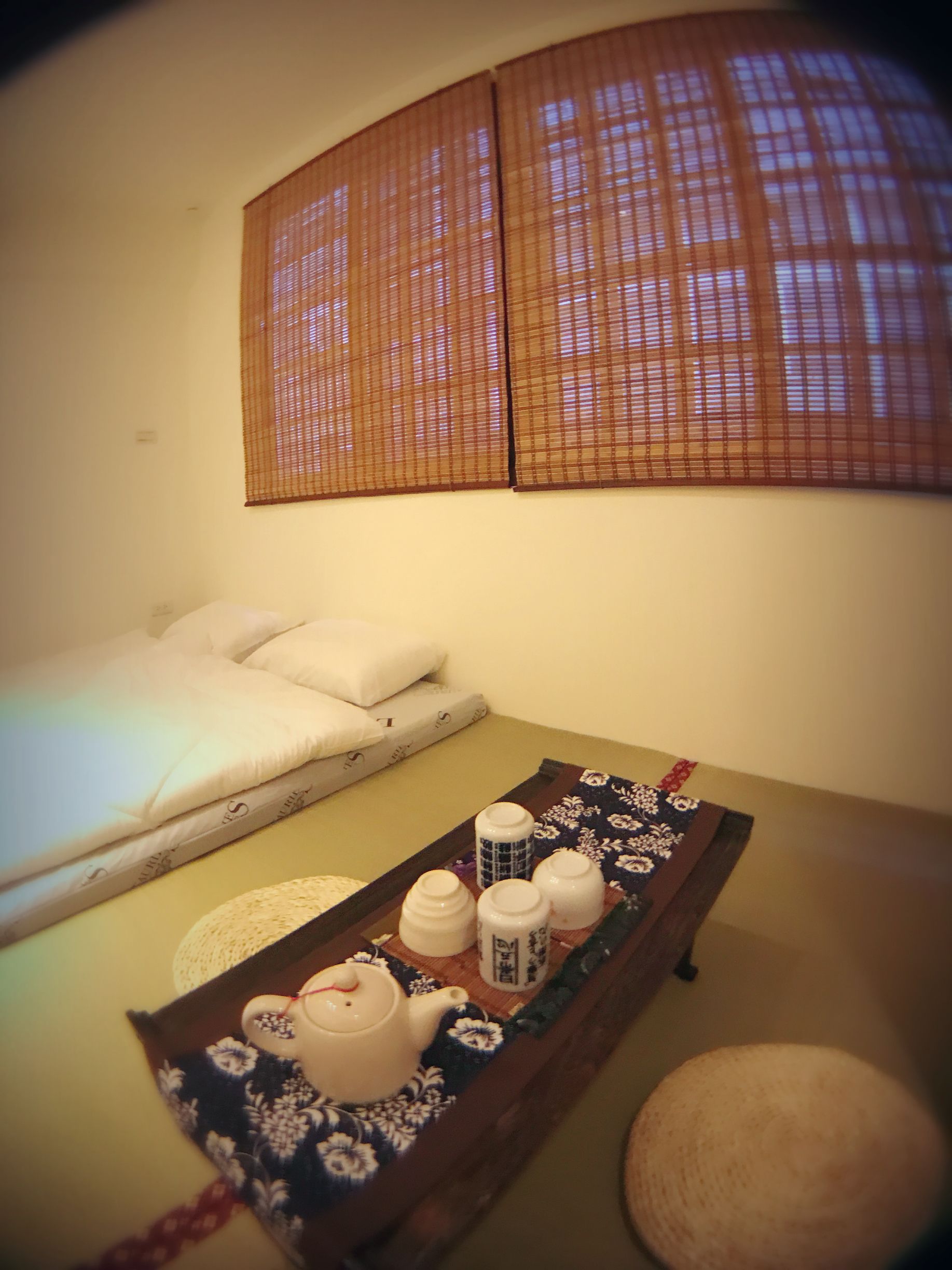 Une pièce avec un lit, une petite table sur laquelle se trouvent une théière et des tasses, et une fenêtre avec des stores.