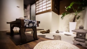 Um quarto japonês tradicional com tatames, uma mesa de madeira baixa com um tecido estampado, um banquinho de fibra natural e um vaso de planta.