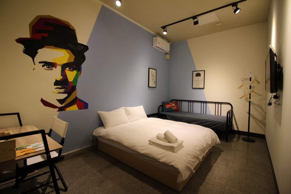 A bedroom with a bed, a desk, a chair, a wall with a colorful mural, and a clothes drying rack.