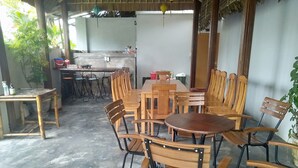 Un comedor con una larga mesa de madera y varias sillas, una pequeña cocina con refrigerador y fregadero, y piso de concreto.