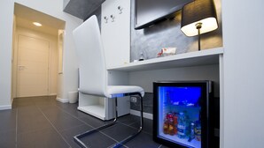 A compact hotel room with a white chair, a small refrigerator with drinks, a TV, and a lamp. - Grey&White B&B (Rome)