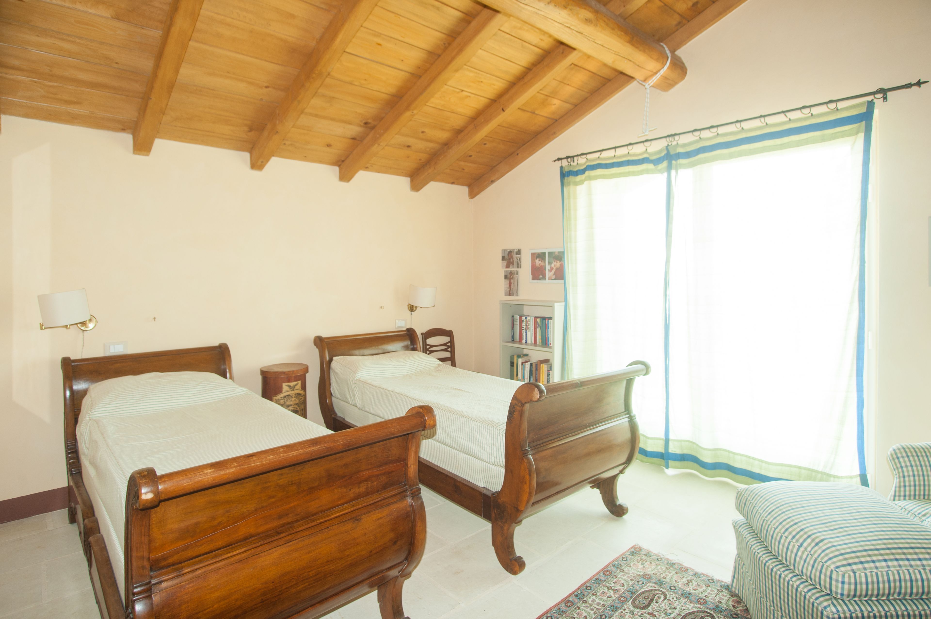 A room with two wooden beds, a small bedside table, a lamp, a bookshelf, and a window with curtains.
