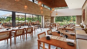 A spacious dining area with wooden tables and chairs, large windows offering views of greenery, and hanging pendant lights. - Woods at Sasan (Talala)