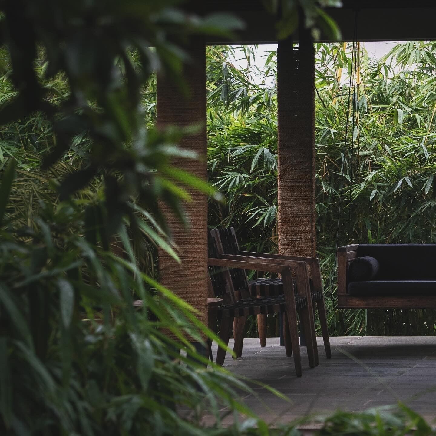 A covered outdoor seating area with wooden chairs and a bench, surrounded by dense green foliage.