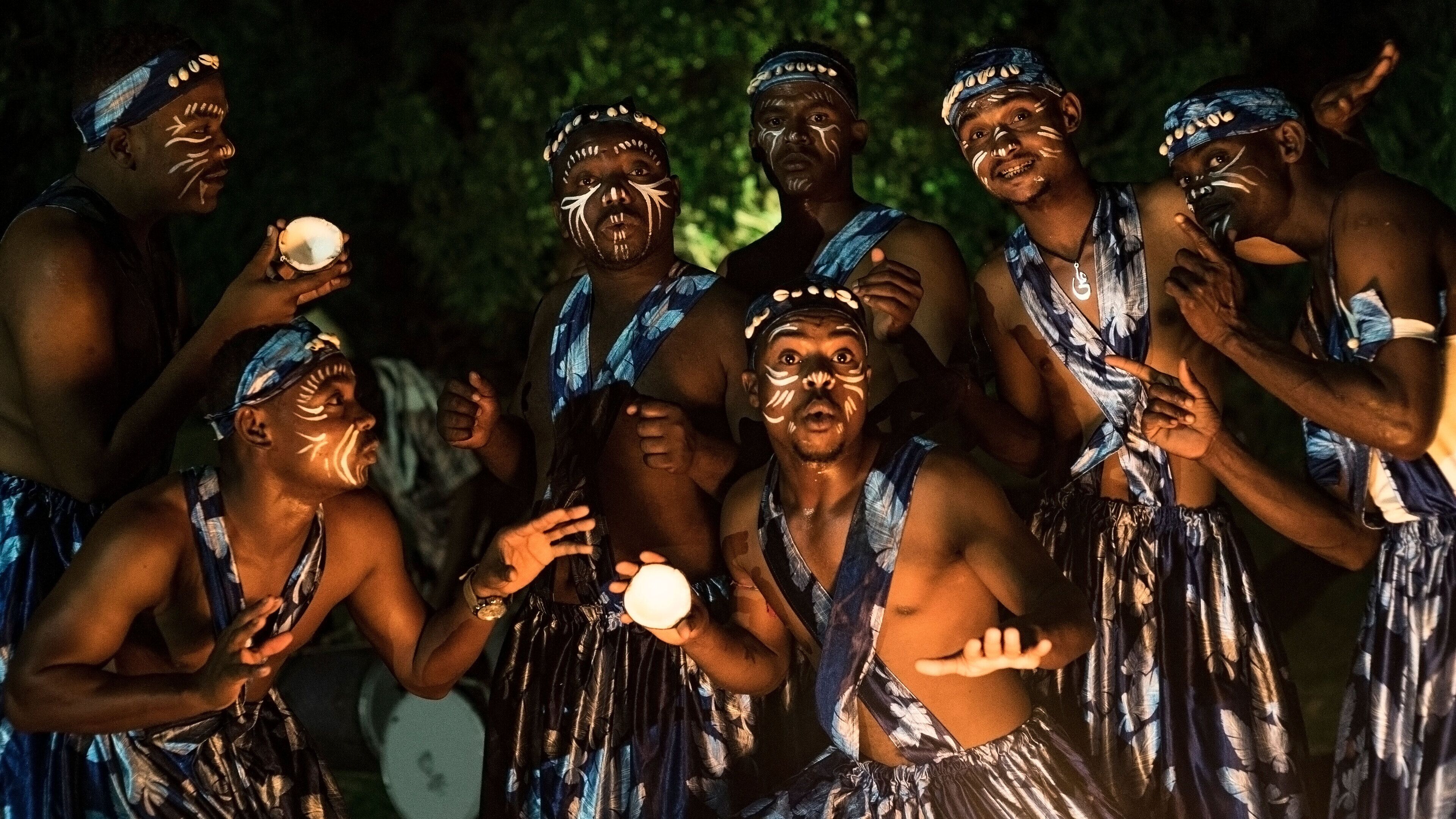 A group of people in traditional attire, some holding objects, in a dimly lit setting.