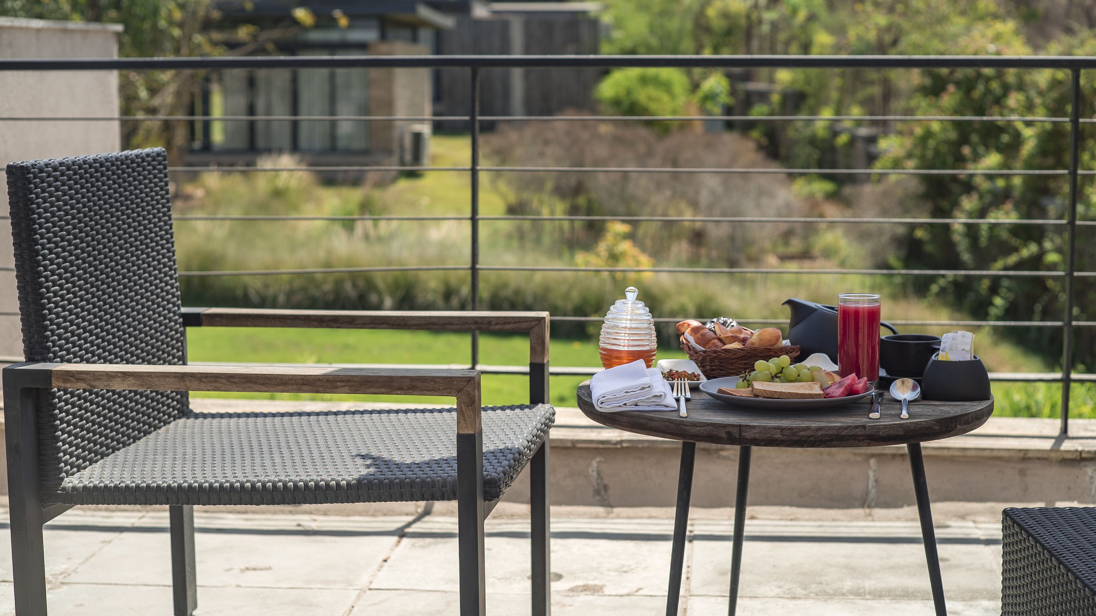 A balcony with wicker chairs, a table with a breakfast spread including bread, fruit, and a drink, and a view of greenery and buildings in the background.