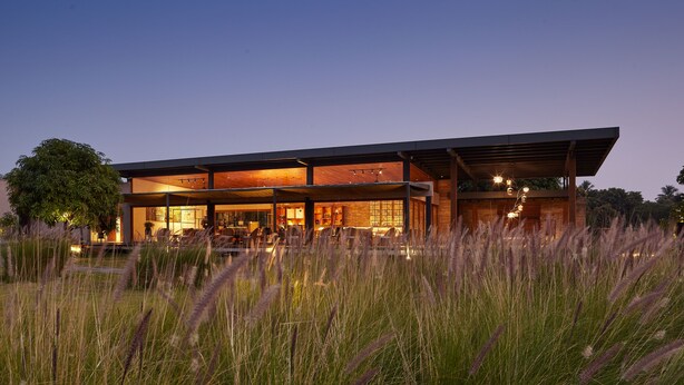 A modern house with a spacious patio area, large windows, and a well-lit interior. - Woods at Sasan (Talala)