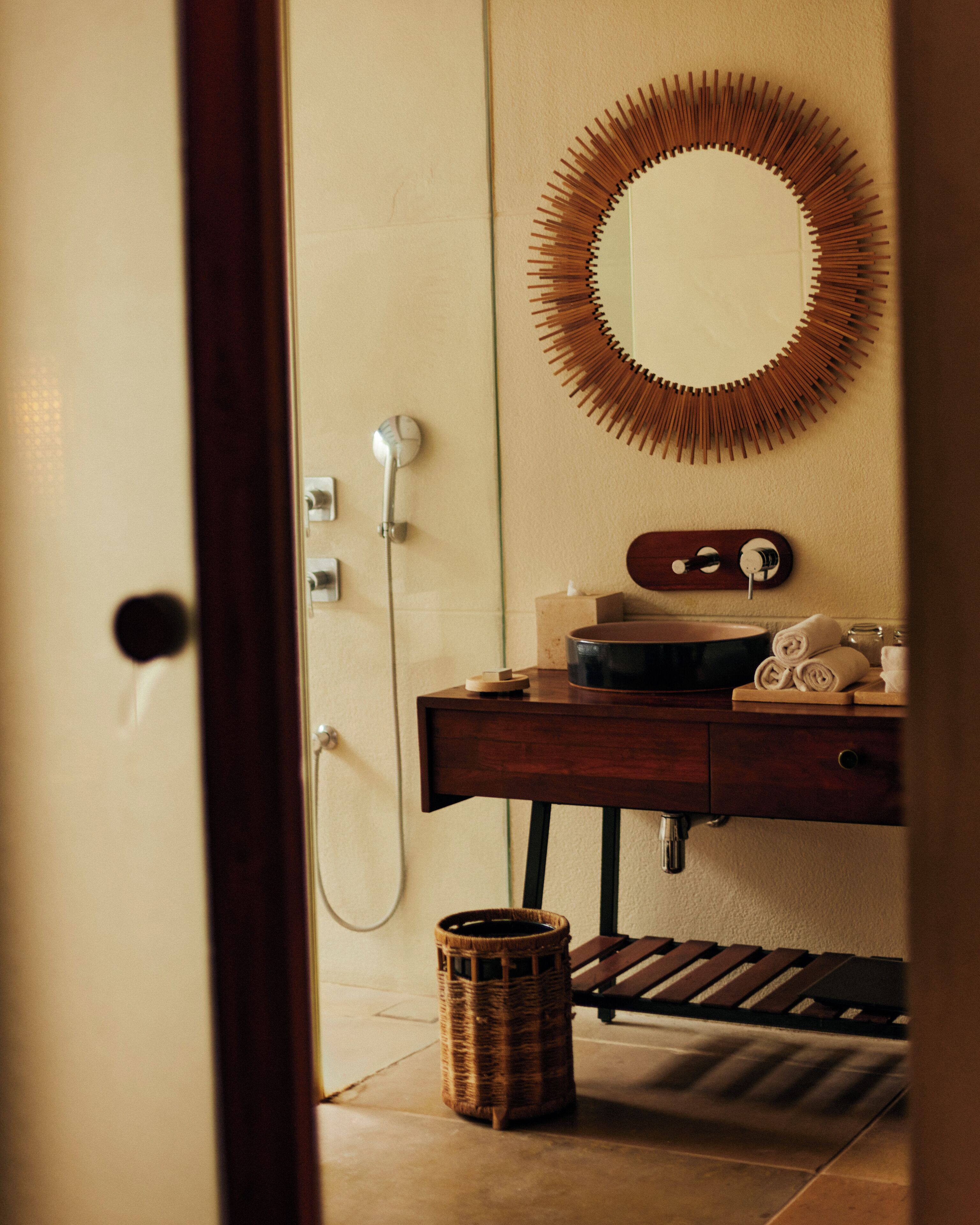 A bathroom with a wooden vanity, a round mirror with a wooden frame, a shower, and a wicker basket on the floor.