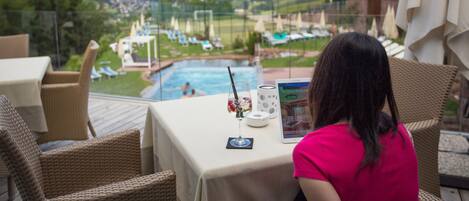 A terrace with wicker furniture, a table with a glass, a tablet, and a view of a pool and mountains.