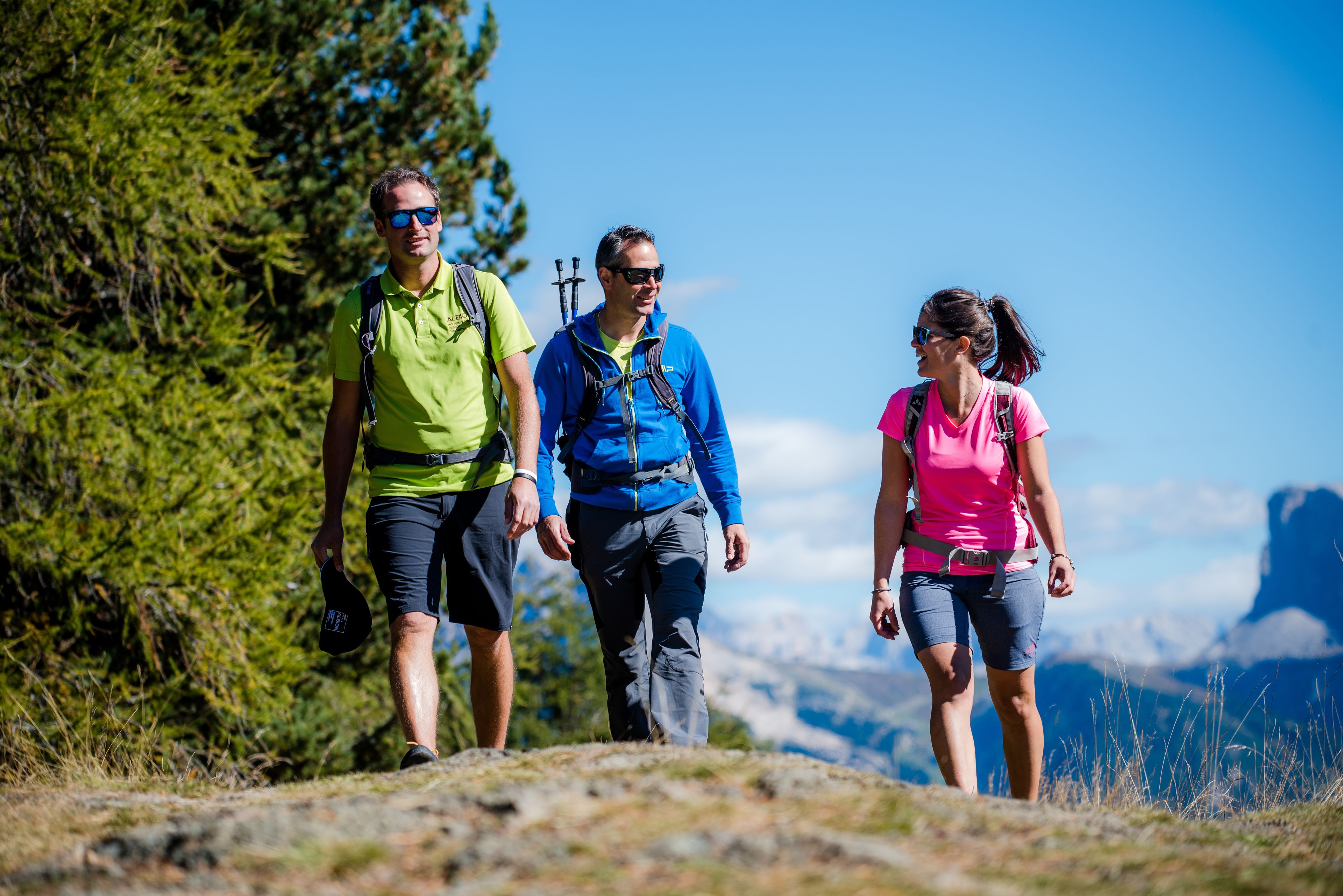 Tres personas caminando por un sendero de montaña, con cielo despejado y un bosque al fondo.